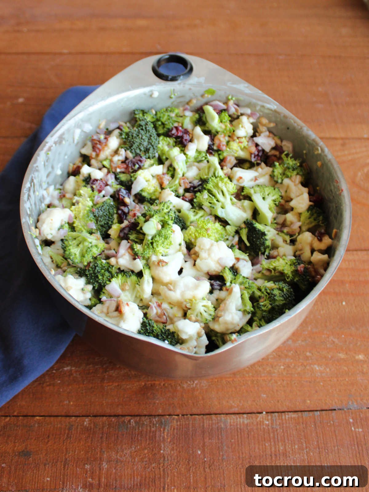 A large mixing bowl filled with freshly prepared broccoli salad, showcasing a delightful blend of crisp broccoli and cauliflower florets, finely diced red onion, crunchy sunflower seeds, savory crumbled bacon, and sweet craisins, all glistening in a light, creamy dressing. The ingredients are perfectly coated and ready to be served.