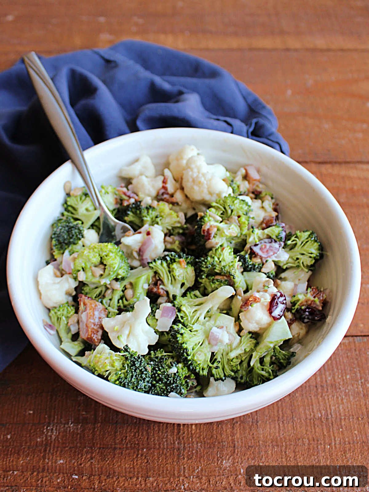 A pristine white serving bowl filled with homemade broccoli salad, perfectly portioned and ready to be enjoyed. A fork rests invitingly in the bowl, highlighting the salad's readiness. The image focuses on the fresh, vibrant textures and colors of the broccoli, cauliflower, and other mix-ins, all coated in the creamy dressing.