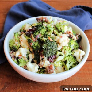A serving bowl filled with crunchy broccoli salad, featuring chunks of fresh broccoli, cauliflower, sweet craisins, savory sunflower seeds, and crispy bacon, all coated in a rich, creamy dressing.