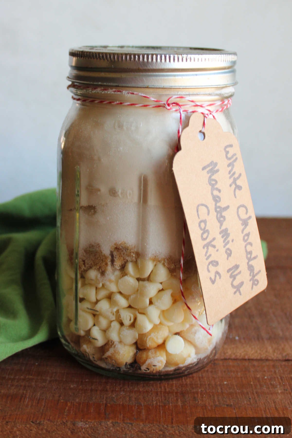 Quart jar filled with layers of macadamia nuts, white chocolate chips, brown sugar, white sugar, and flour mixed with salt, baking powder and baking soda to make white chocolate macadamia nut cookies. 
