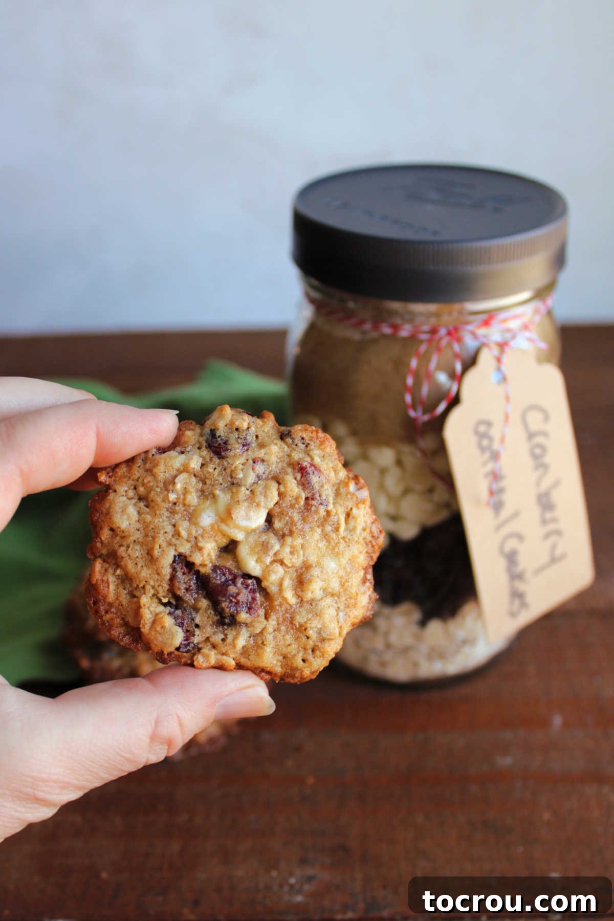 Homemade Cranberry Oatmeal Cookie Hand holding a cranberry white chocolate oatmeal cookie next to a jar of homemade cookie mix. A freshly baked cookie is shown, highlighting the delicious result of the easy-to-use mix, inviting you to bake your own.