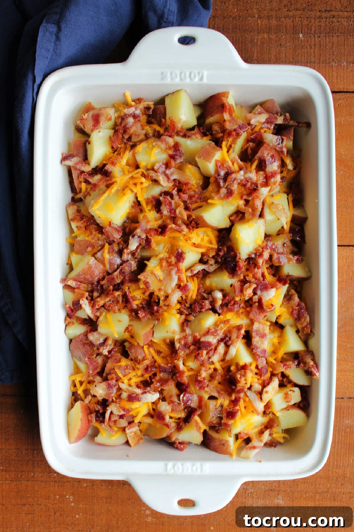 Casserole dish with cooked potatoes topped with shredded cheddar cheese and chopped cooked bacon ready to go in the oven.