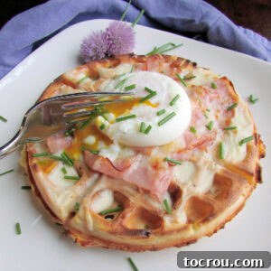 Fork breaking into a runny poached egg nestled over a ham and cheese waffle topped with creamy bechamel sauce, ham, and melted cheese.
