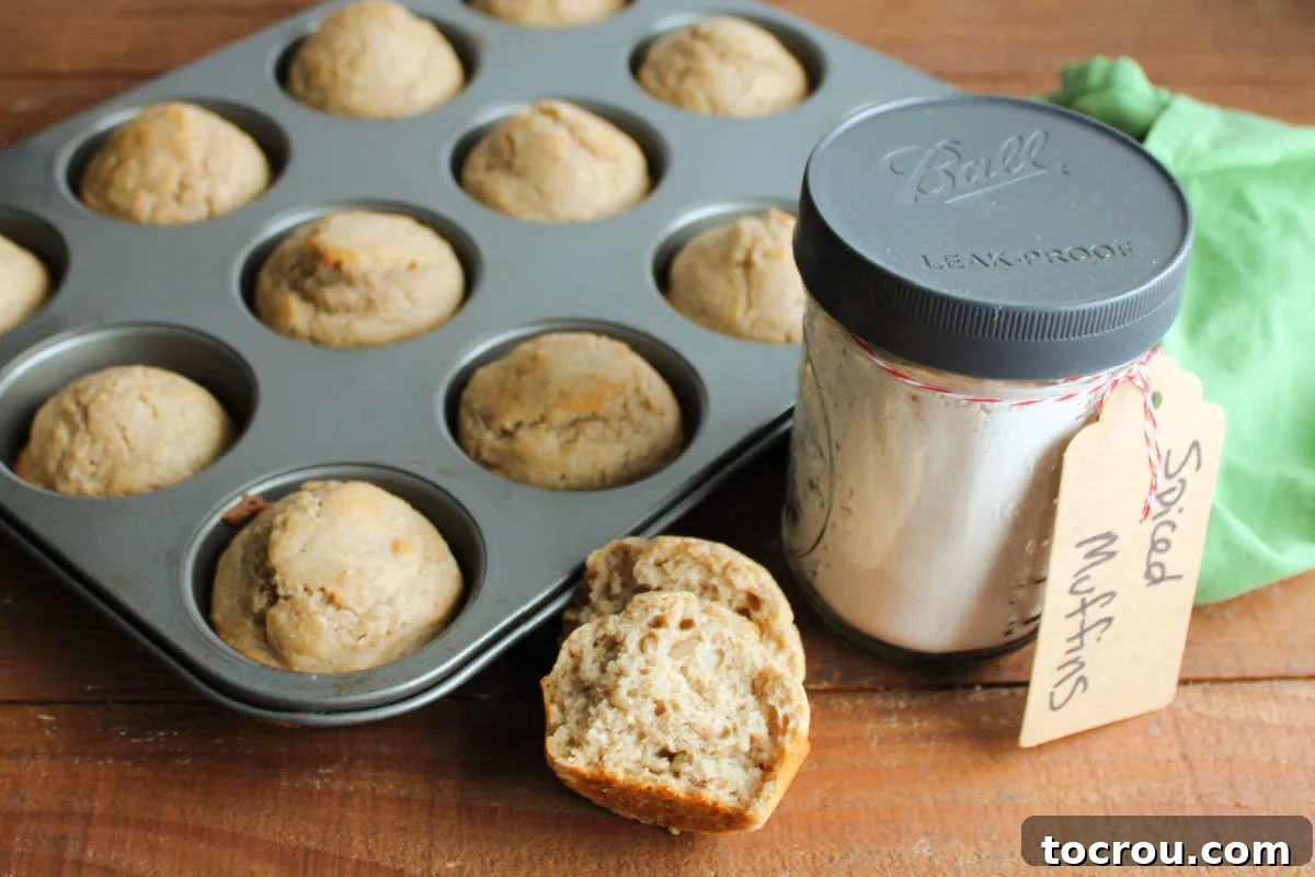 A muffin tin filled with freshly baked spiced muffins, a muffin torn in half revealing its soft interior, and a clear jar of homemade spiced muffin mix, ready to be stored or gifted.