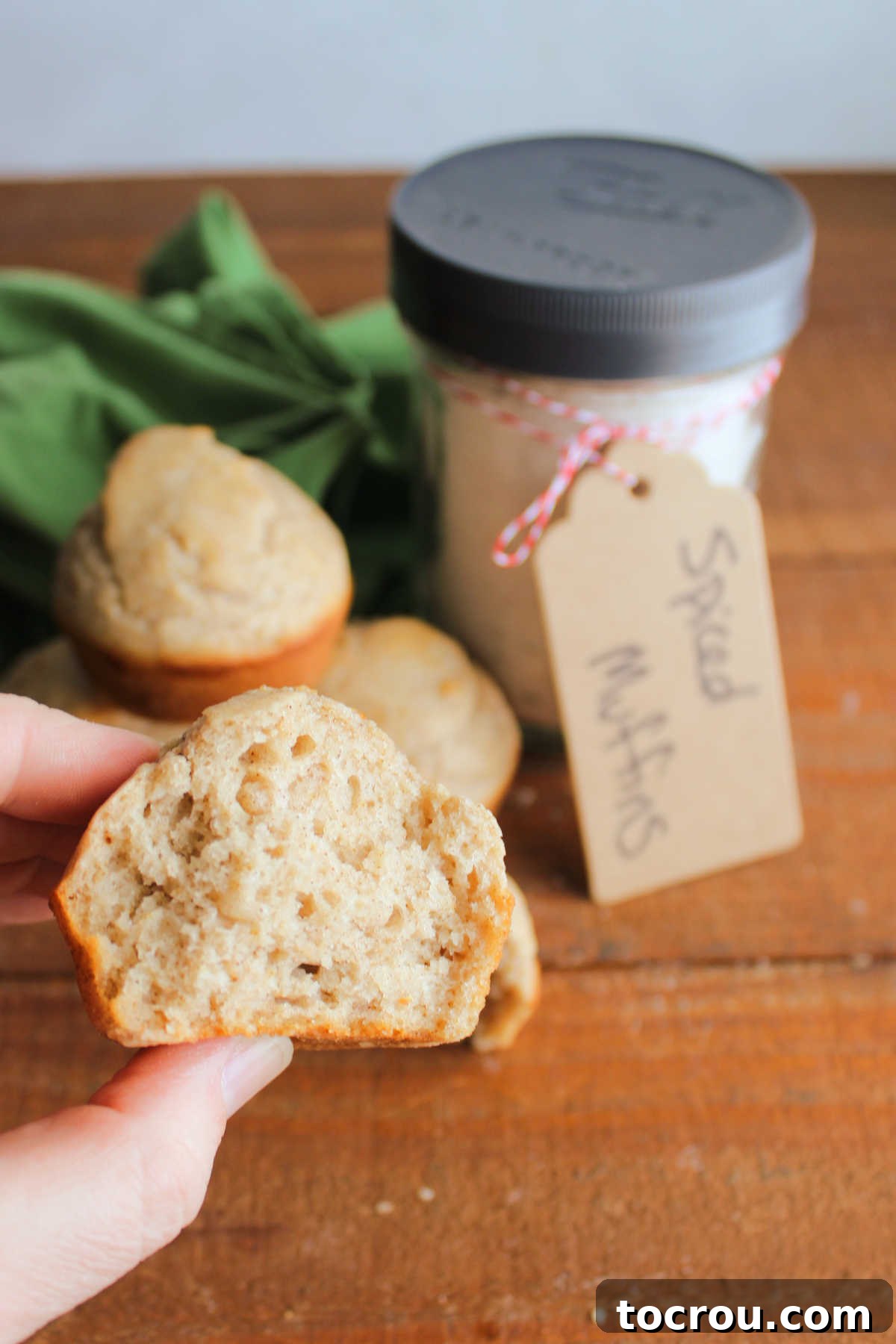 A hand gently holds half of a soft, freshly baked spiced muffin, showcasing its tender texture. In the background, a jar of homemade muffin mix and a collection of baked muffins complete the inviting scene.