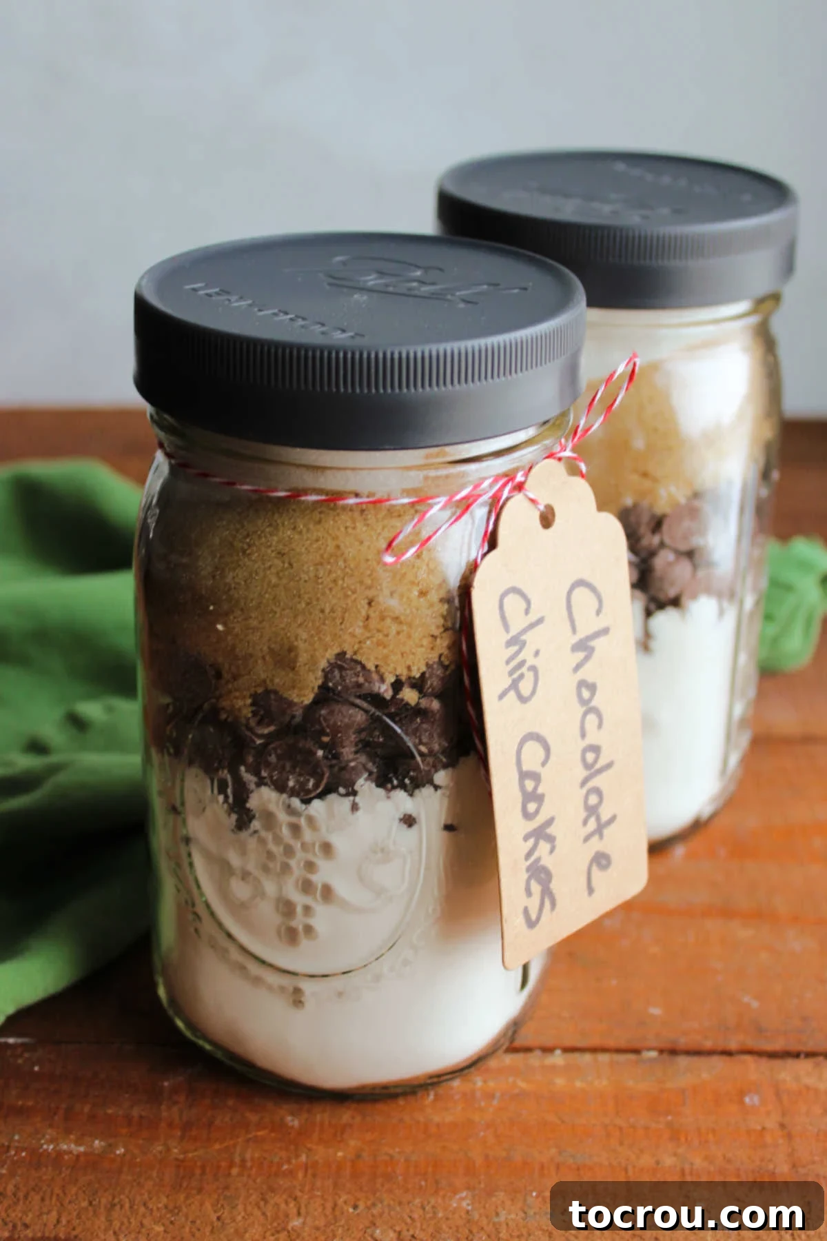 DIY Chocolate Chip Cookie Jar Mix 3 Two glass quart jars filled with the dry ingredients to make chocolate chip cookies including a flour mixture, chocolate chips, brown sugar, and granulated sugar with a tag tied to the jar.