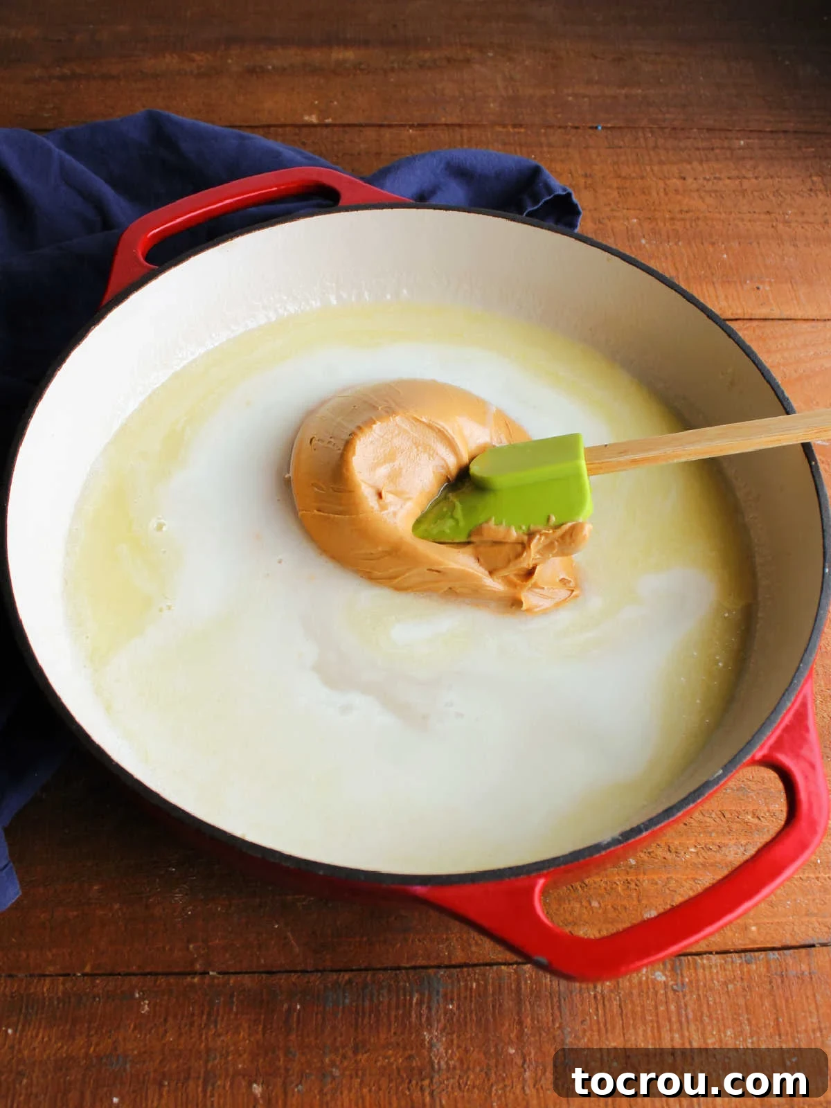 Smooth, creamy peanut butter being stirred into a warm, shimmering mixture of sugar, milk, and melted butter in a saucepan.
