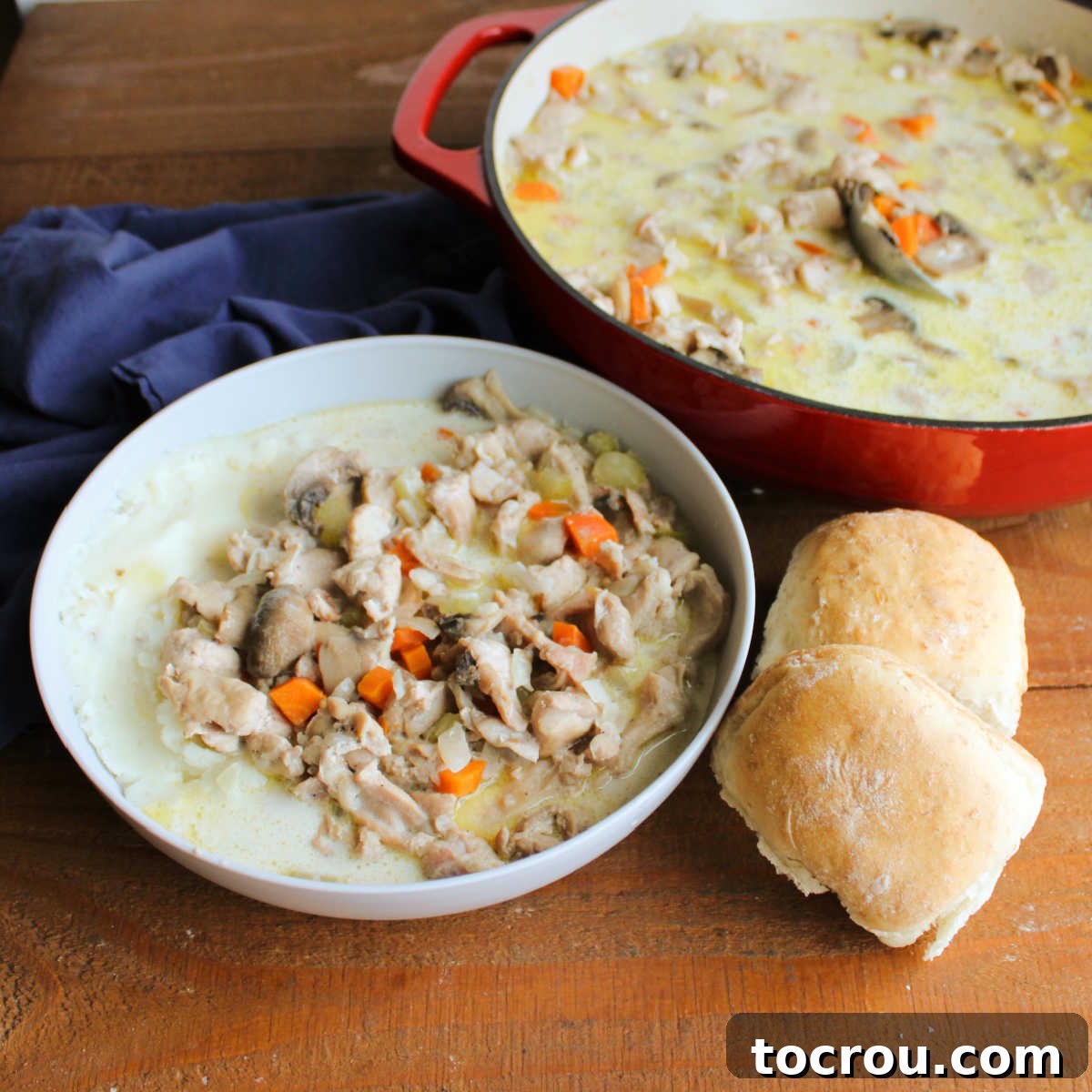 Bowl of creamy German rabbit stew next to some homemade dinner rolls with the pan of more German style rabbit stew in the background, inviting diners to a hearty meal.