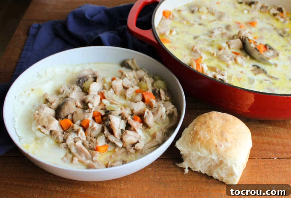 A hearty bowl of rabbit stew, featuring a luxuriously creamy broth studded with tender chunks of rabbit, vibrant vegetables, and earthy mushrooms. The stew is elegantly served over a bed of fluffy mashed potatoes, with a warm, homemade dinner roll nestled invitingly nearby, promising a truly satisfying meal.