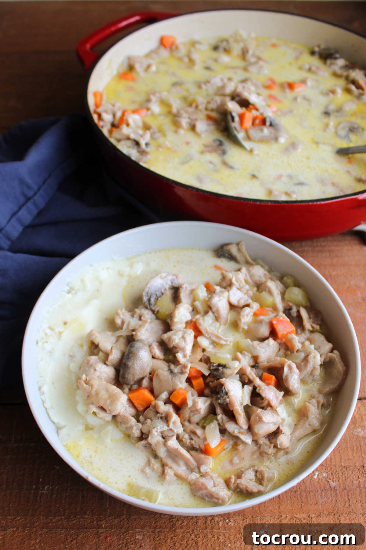 A beautifully presented bowl of creamy rabbit stew, generously spooned over a bed of smooth mashed potatoes. In the soft-focused background, an enameled cast iron braiser holds more of the simmering stew, suggesting a feast ready to be enjoyed.