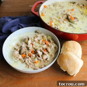 Bowl of creamy rabbit stew next to some homemade dinner rolls with the pan of more German style rabbit stew in the background.