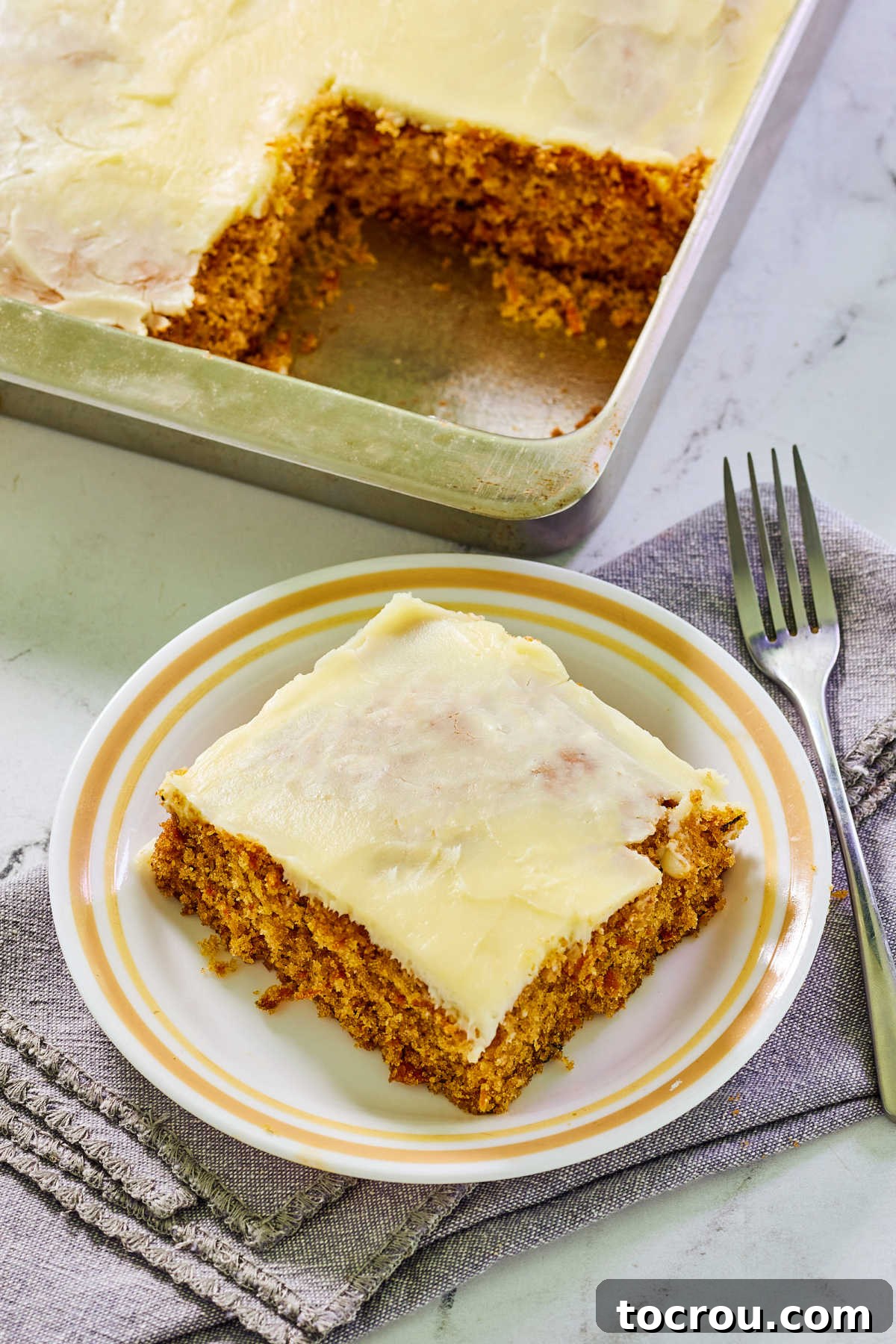 Irresistible Homemade Carrot Cake 9 Plate with a piece of carrot cake topped with cream cheese icing next to pan with remaining cake inside.