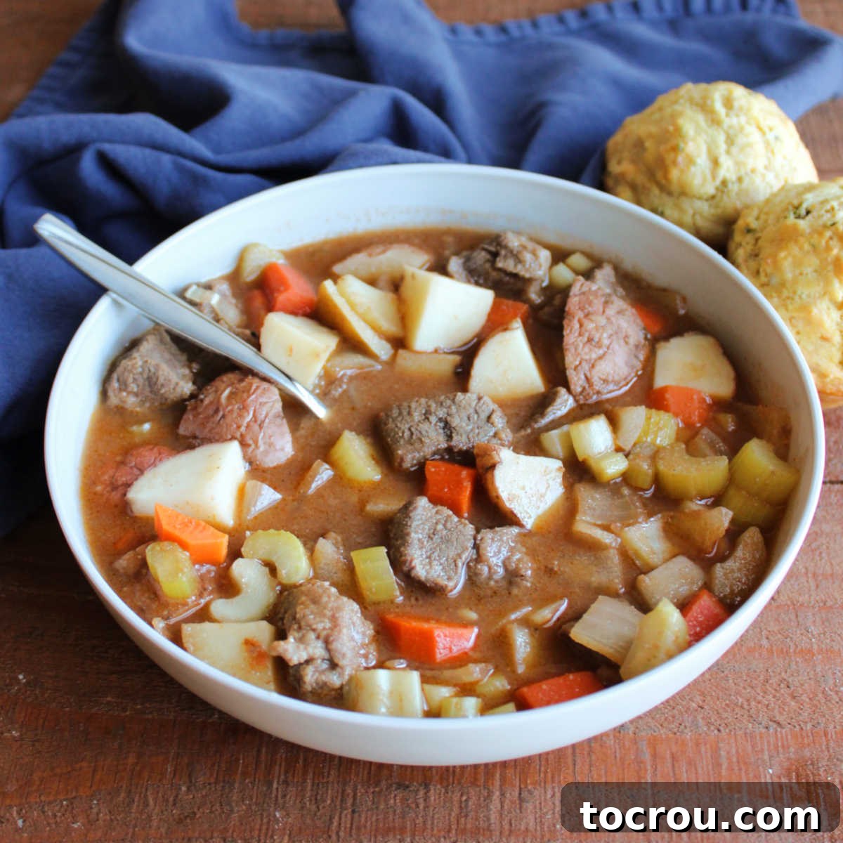 Hearty Venison Stew with Muffins A steaming bowl of venison stew, brimming with tender chunks of deer meat, potatoes, carrots, onions, and celery, served in a rich, dark broth. Savory Parmesan herb muffins are artfully placed nearby, ready for dipping.