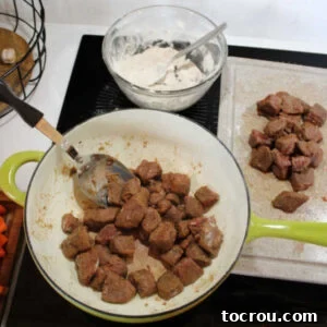 Browning coated venison in batches with some browned meat on a cutting board, some in the pot, and the bowl of extra flour nearby.