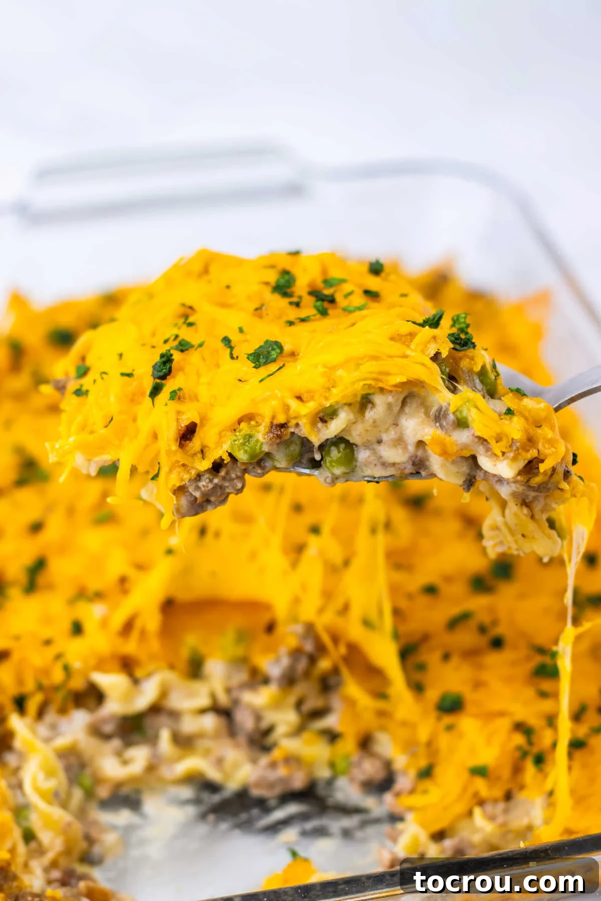 A serving spoon lifting a generous portion of hot Amish Country Casserole out of a baking dish, showcasing the abundant melted cheese, ground beef, egg noodles, and peas.
