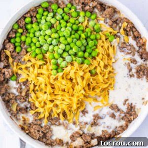 Skillet filled with ground beef and onion in sauce made from milk and cream of mushroom soup with uncooked egg noodles and peas on top.