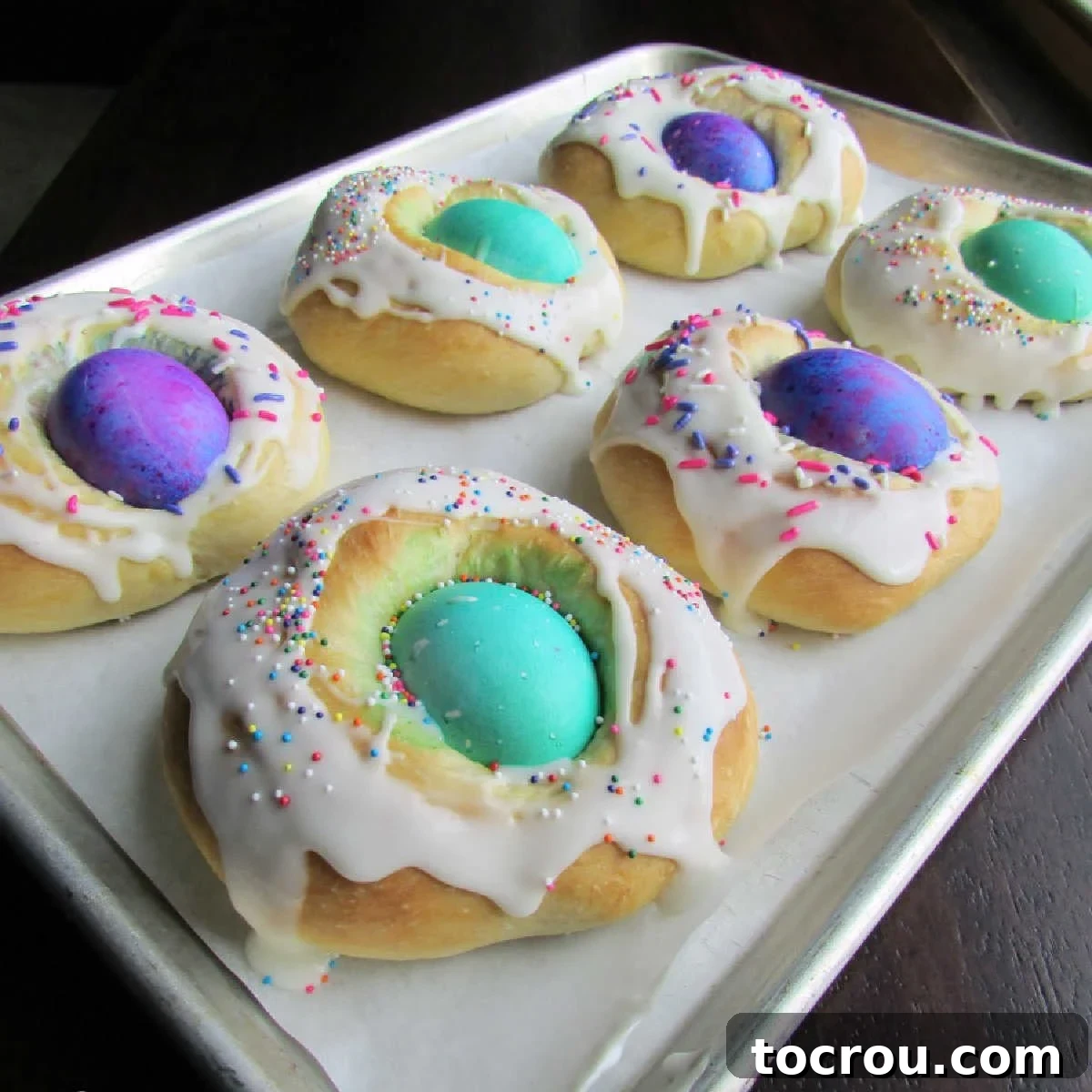 Baking tray with Easter bread wreaths with dyed hard boiled eggs in the center and a powdered sugar icing and sprinkles over the surrounding bread.