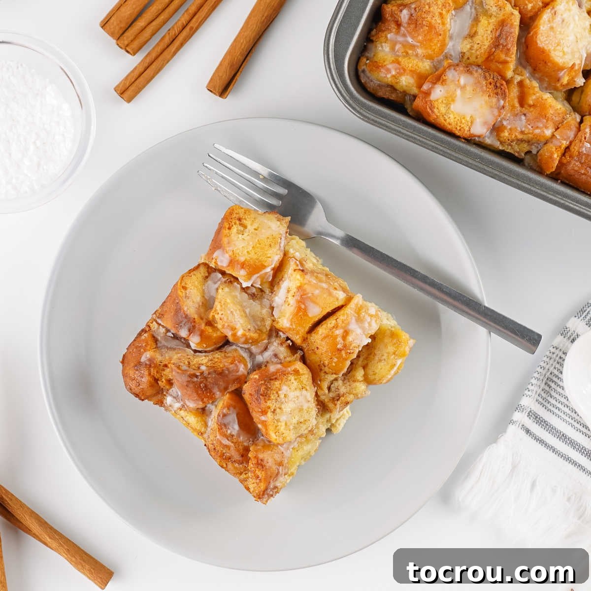 A square portion of golden-brown donut bread pudding on a dessert plate, with a larger baking pan of the pudding and cinnamon sticks in the background. The rich texture and inviting glaze hint at its deliciousness.