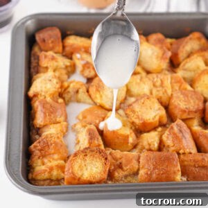 Spoon drizzling powdered sugar glaze over pan of hot donut bread pudding.