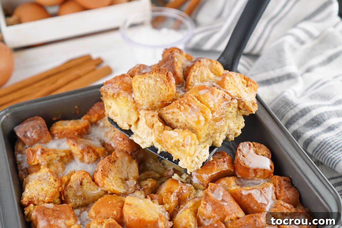 A spatula lifting a glistening piece of freshly baked donut bread pudding from a baking pan, showcasing its golden crust and soft interior.
