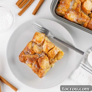 square of donut bread pudding on dessert plate with baking pan with more bread pudding and some cinnamon sticks nearby.