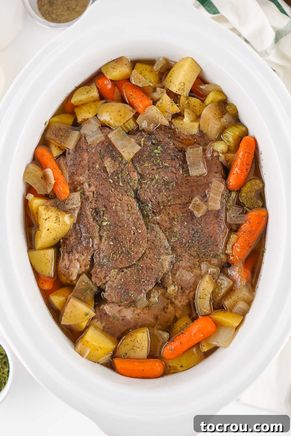 Crockpot filled with cooked pot roast with tender cooked chuck roast surrounded by cooked carrots, chunks of potato, celery, and onions in broth.