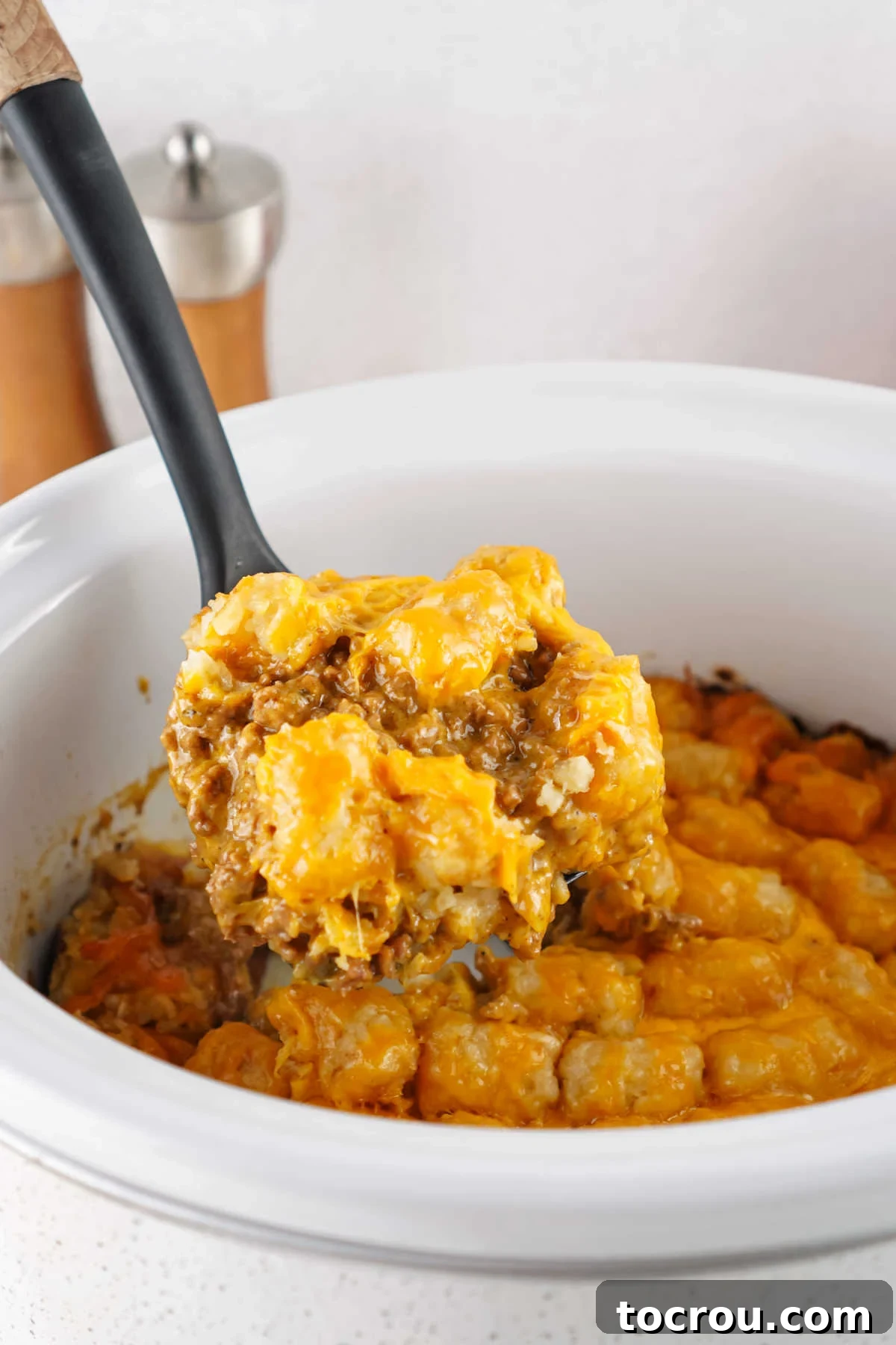 Serving spoon getting a helping of taco tater tot casserole out of a crockpot showing cheesy seasoned ground beef topped with tater tots and melted cheese. 