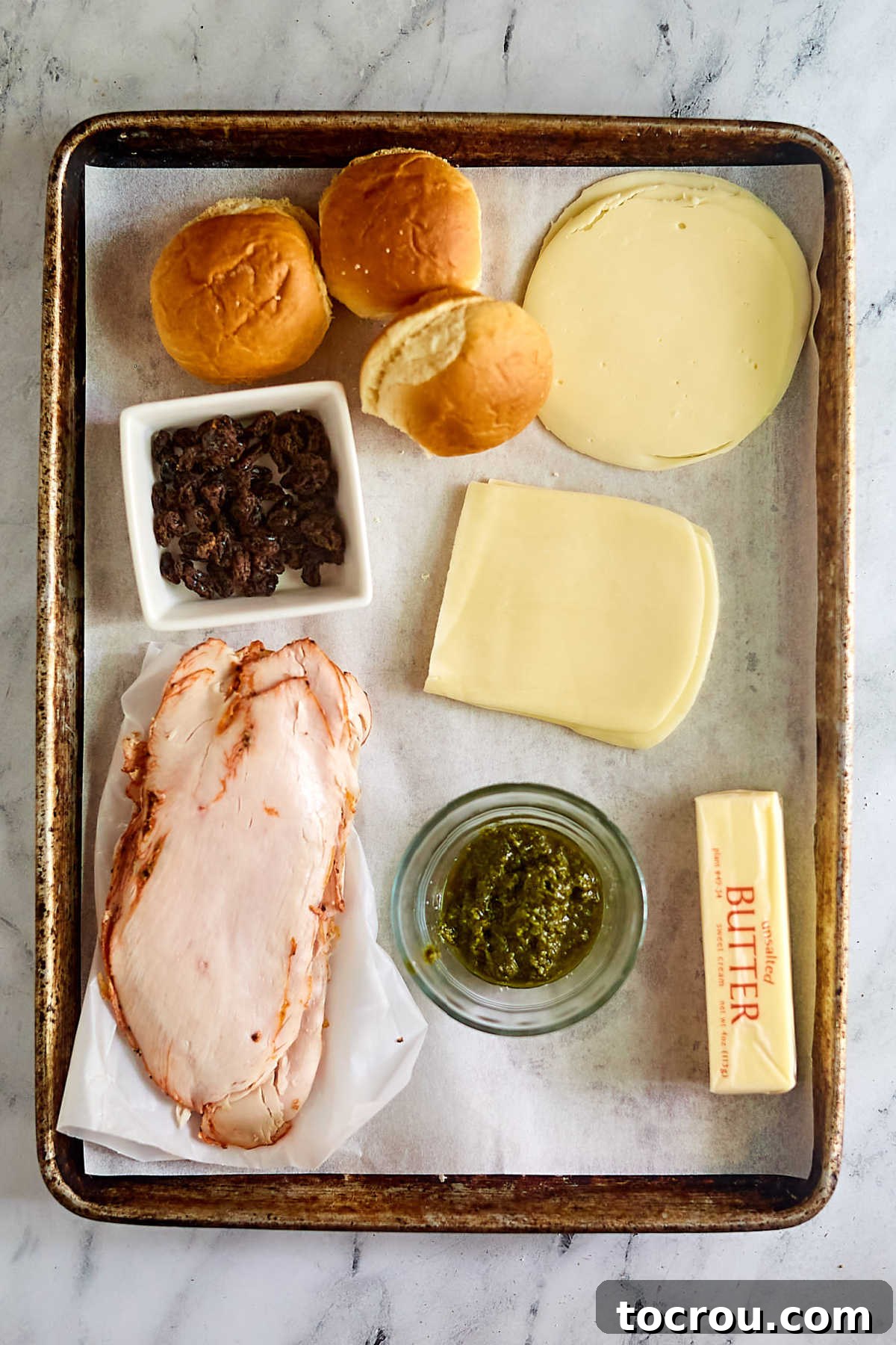 Essential ingredients for turkey grilled cheese sliders laid out, including fresh rolls, assorted cheeses, vibrant pesto, thinly sliced turkey, rich butter, and dried cranberries.