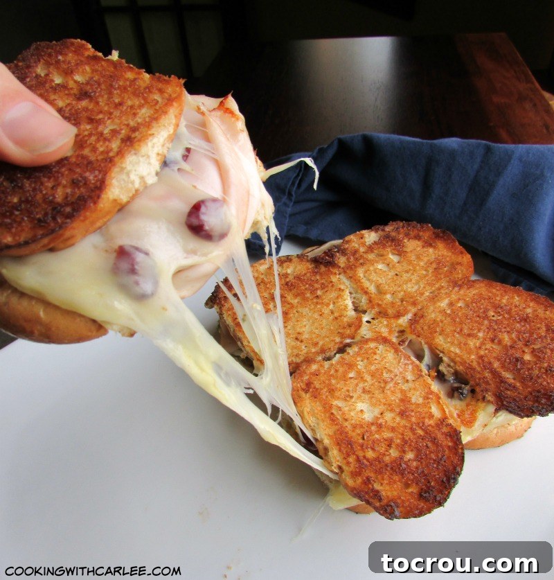 A hand reaching for a turkey grilled cheese slider, with strings of melted cheese stretching and a glimpse of the vibrant craisins within.
