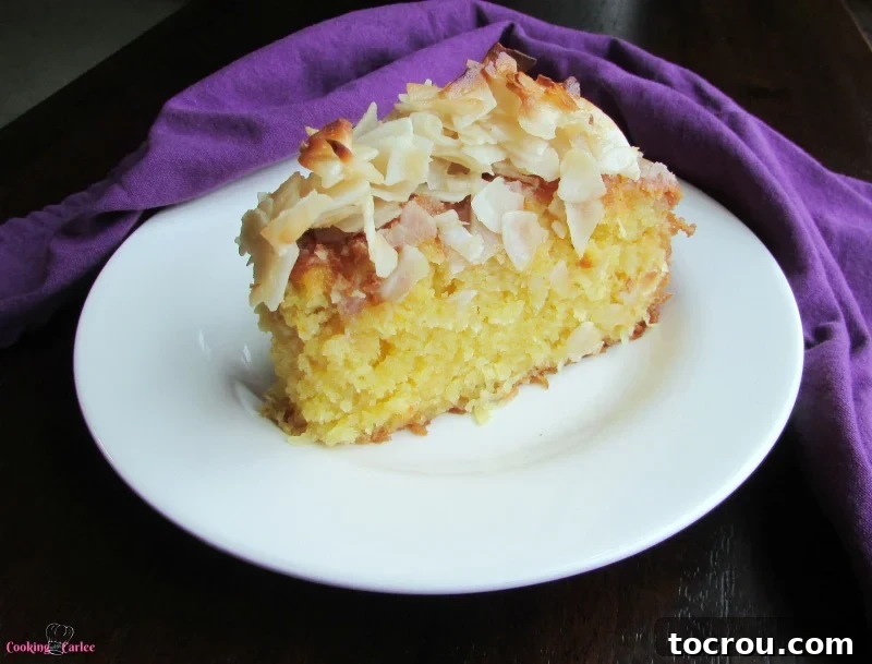 A generous slice of rich, yellow-hued Greek coconut cake, visibly soaked in sweet syrup and adorned with large, perfectly browned flakes of coconut on top.