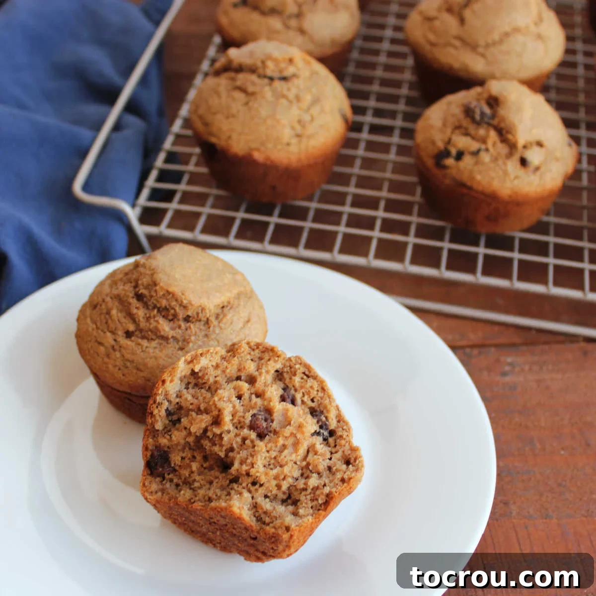 Half of a bran muffin on a plate showing its airy interior dotted with raisins with more freshly baked homemade bran muffins in the background. The muffin is golden brown with a slight dome, and the interior reveals a soft, porous texture with visible specks of bran and plump raisins.