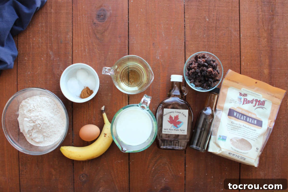 Ingredients including whole wheat flour, baking powder, baking soda, salt, cinnamon, egg, banana, milk, oil, maple syrup, raisins, vanilla and wheat bran are meticulously arranged on a countertop, ready to be transformed into delicious bran muffins. Each item is clearly visible and pre-measured.