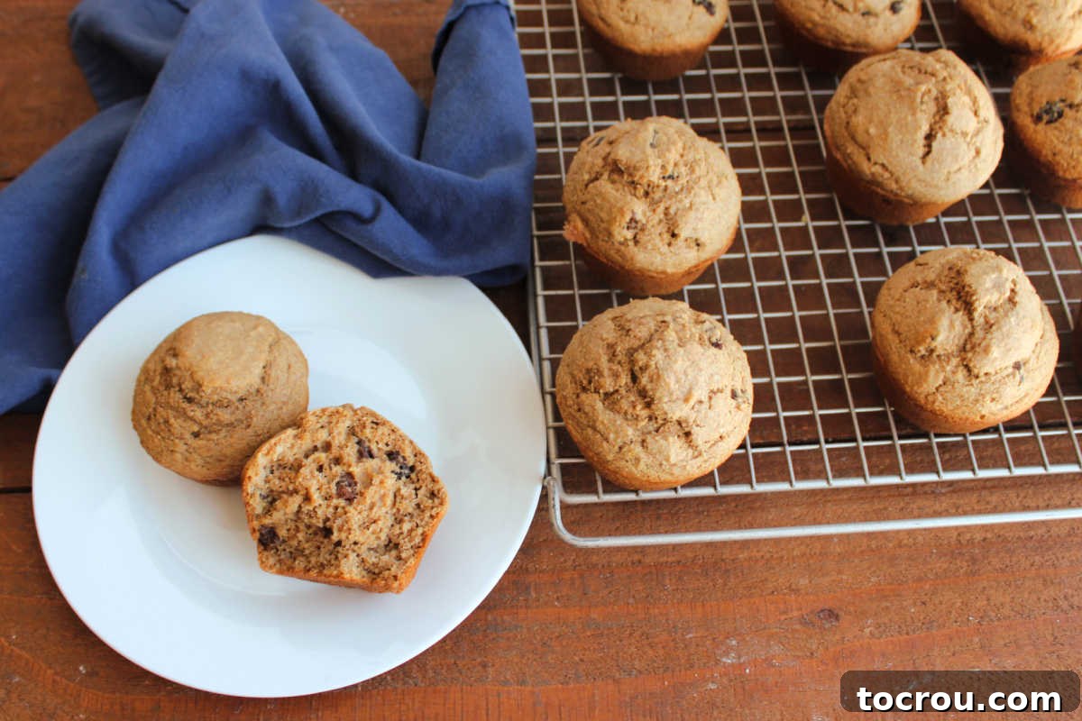 Two perfectly baked homemade bran muffins are elegantly displayed on a small white plate, their tops golden and inviting. In the soft-focus background, a wire cooling rack holds a generous batch of additional freshly baked bran muffins, promising a delicious spread.