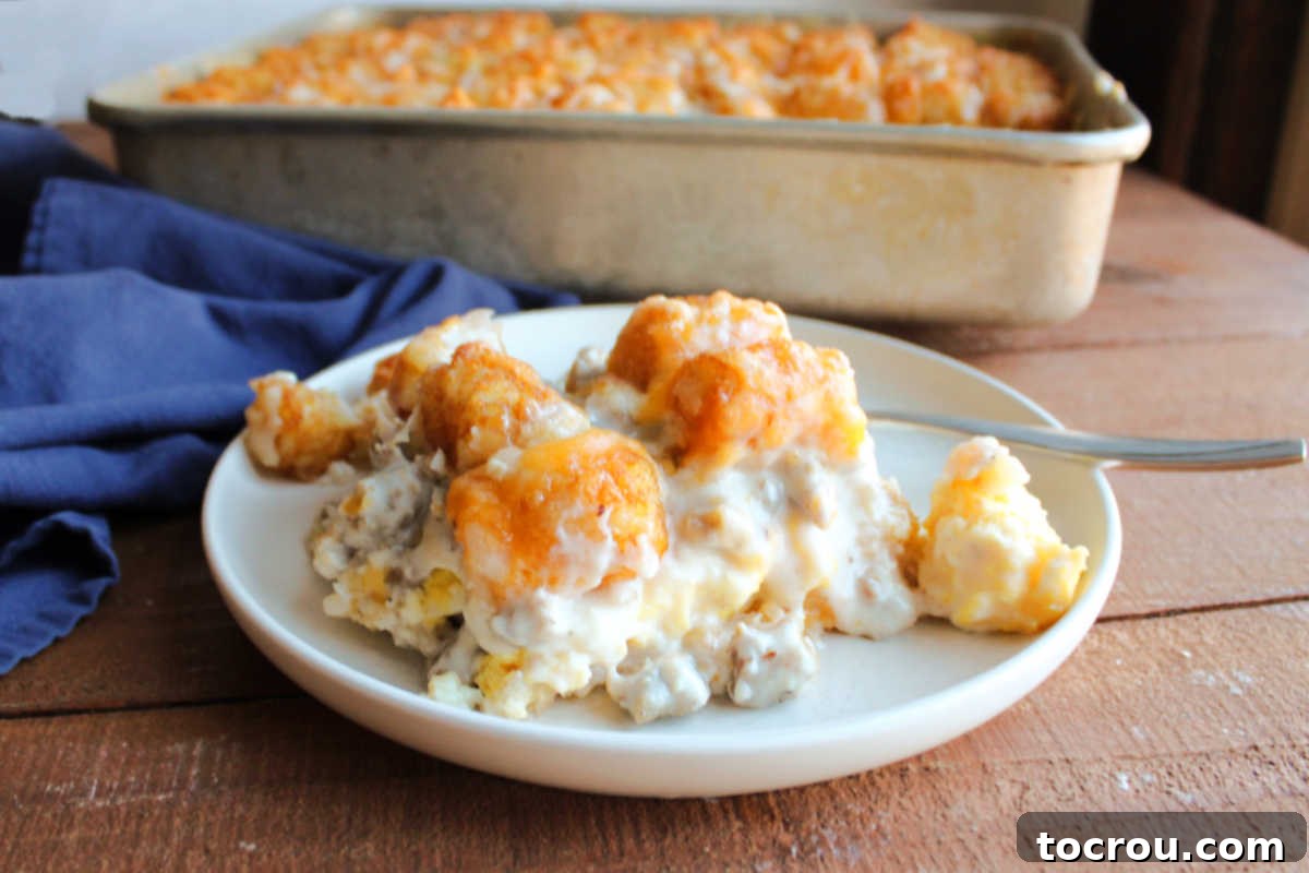Biscuits and Gravy Tater Tot Casserole Plate with a piece of sausage gravy tater tot casserole on it showing the layers of biscuits, eggs, gravy, and tater tots with a fork and the pan of remaining casserole in the background.