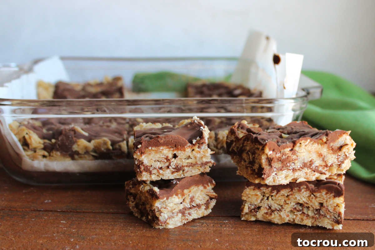 Small, perfectly cut squares of no-bake oatmeal cookie bars, featuring visible layers of oats and chocolate peanut butter, arranged in front of a baking pan containing more uncut bars.