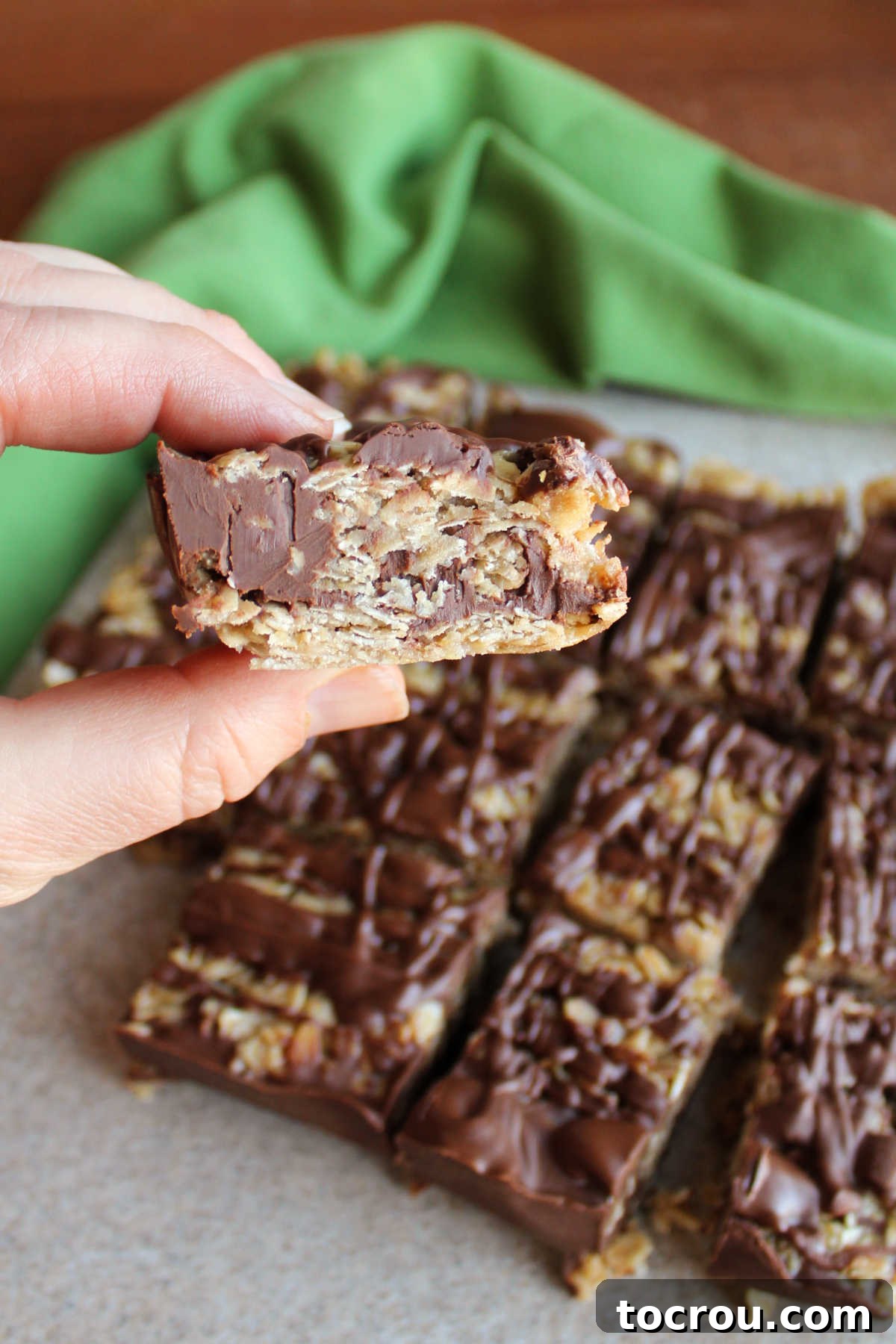 A hand holding a single no-bake oatmeal cookie bar, showing the thick layers of chocolate peanut butter and the oat mixture, with a cutting board of more bars in the blurred background.