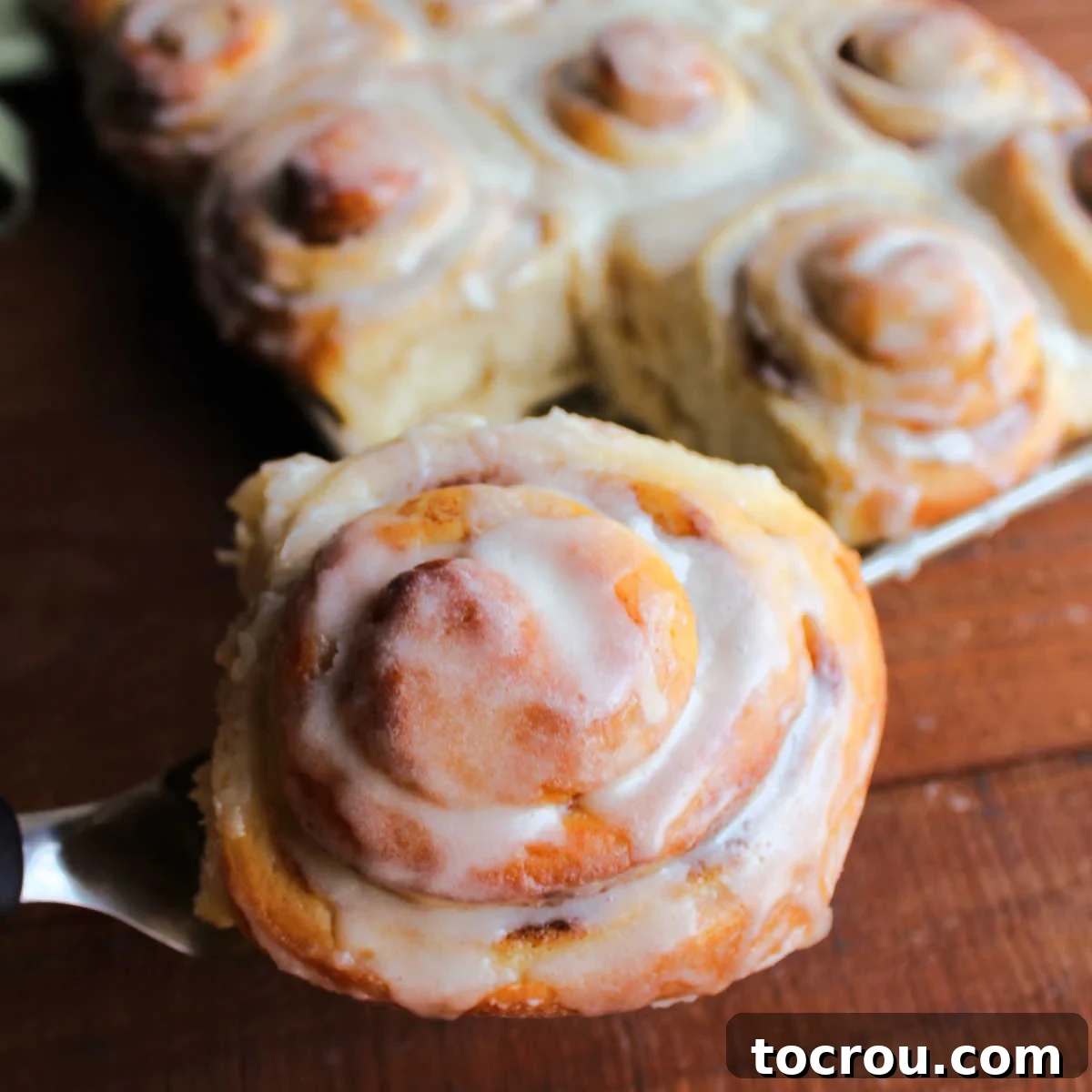Spatula lifting large golden brown potato cinnamon roll topped with vanilla icing out of pan of freshly baked cinnamon rolls.