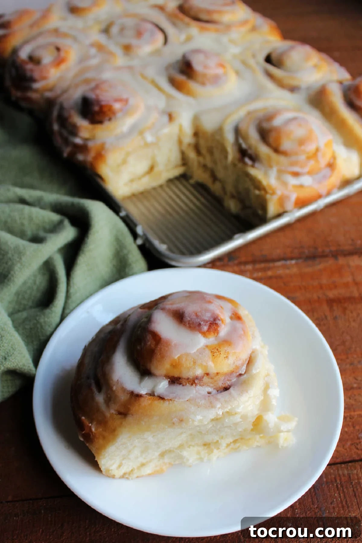 Fluffy cinnamon roll on plate in front of pan of remaining potato cinnamon rolls.
