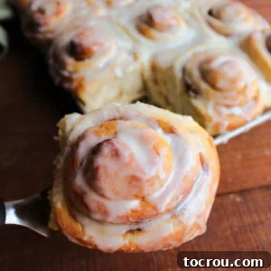 Spatula lifting large golden brown potato cinnamon roll topped with vanilla icing out of pan of freshly baked cinnamon rolls.