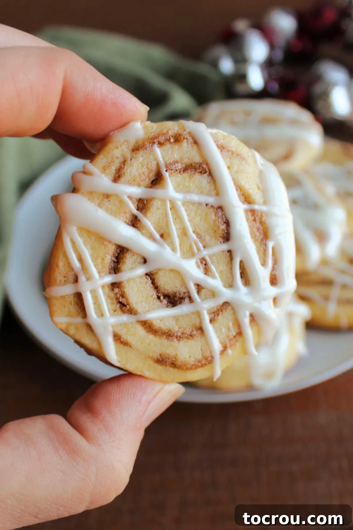 Hand holding a cinnamon roll cookie with swirls of cinnamon filling baked inside and cream cheese icing drizzled over the top.