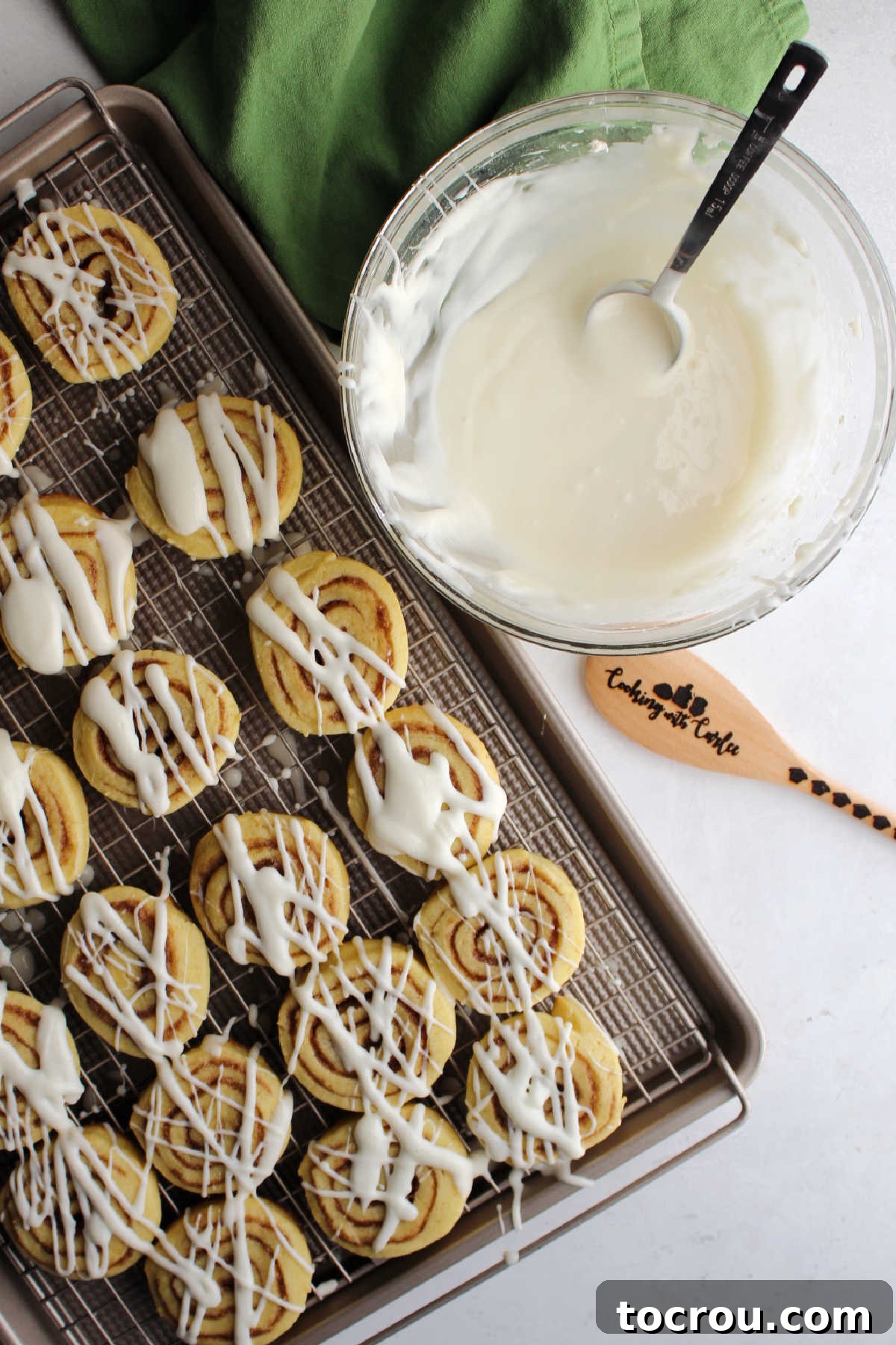 Cinnamon roll cookies with stripes of cream cheese icing next to bowl with more cream cheese icing.