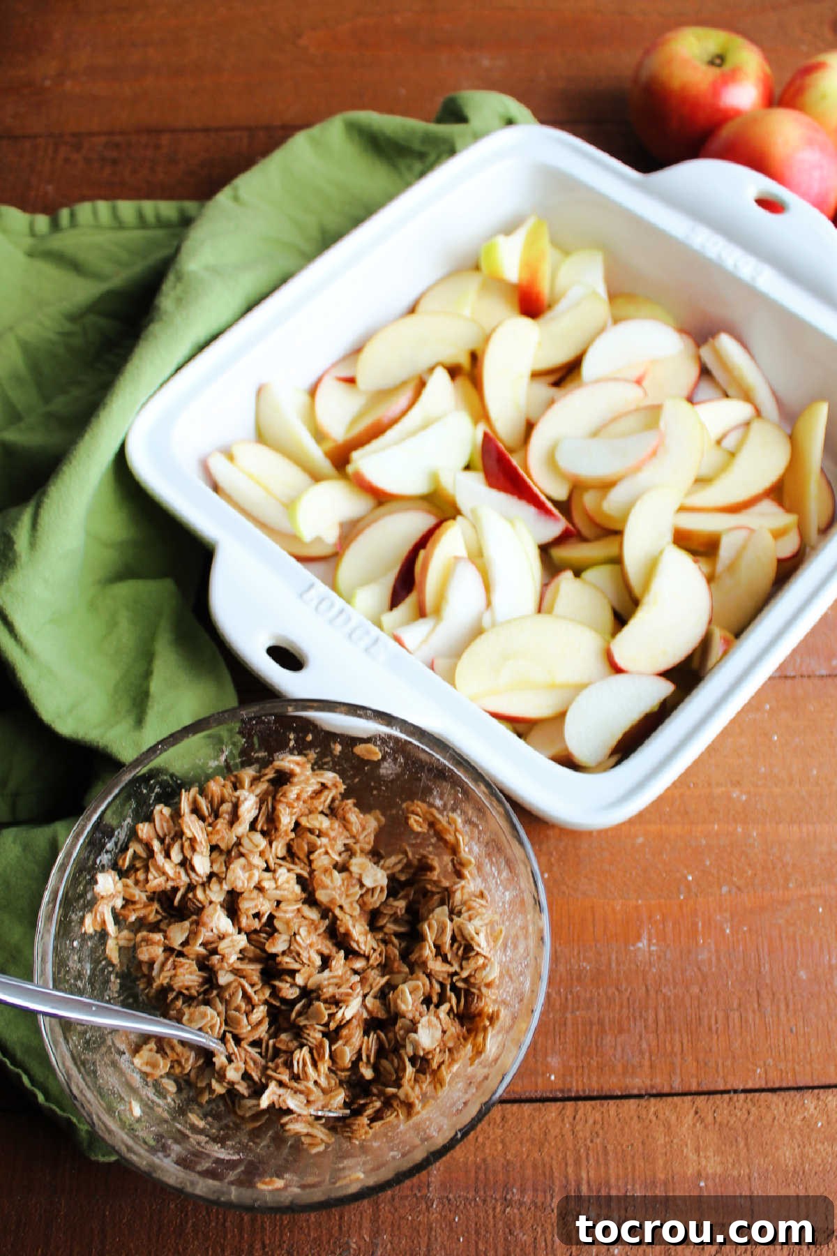 Sliced apples in a square baking dish with a small bowl of oat topping nearby, ready for assembly.