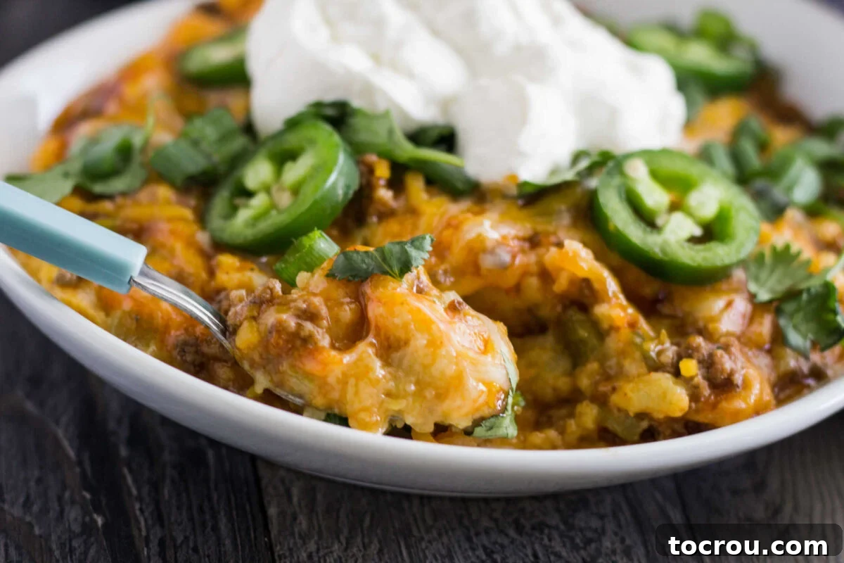 Bite of tamale casserole with soft cornmeal layer topped with enchilada sauce, ground meat and melted cheese garnished with jalapeno slices, cilantro, and sour cream. 