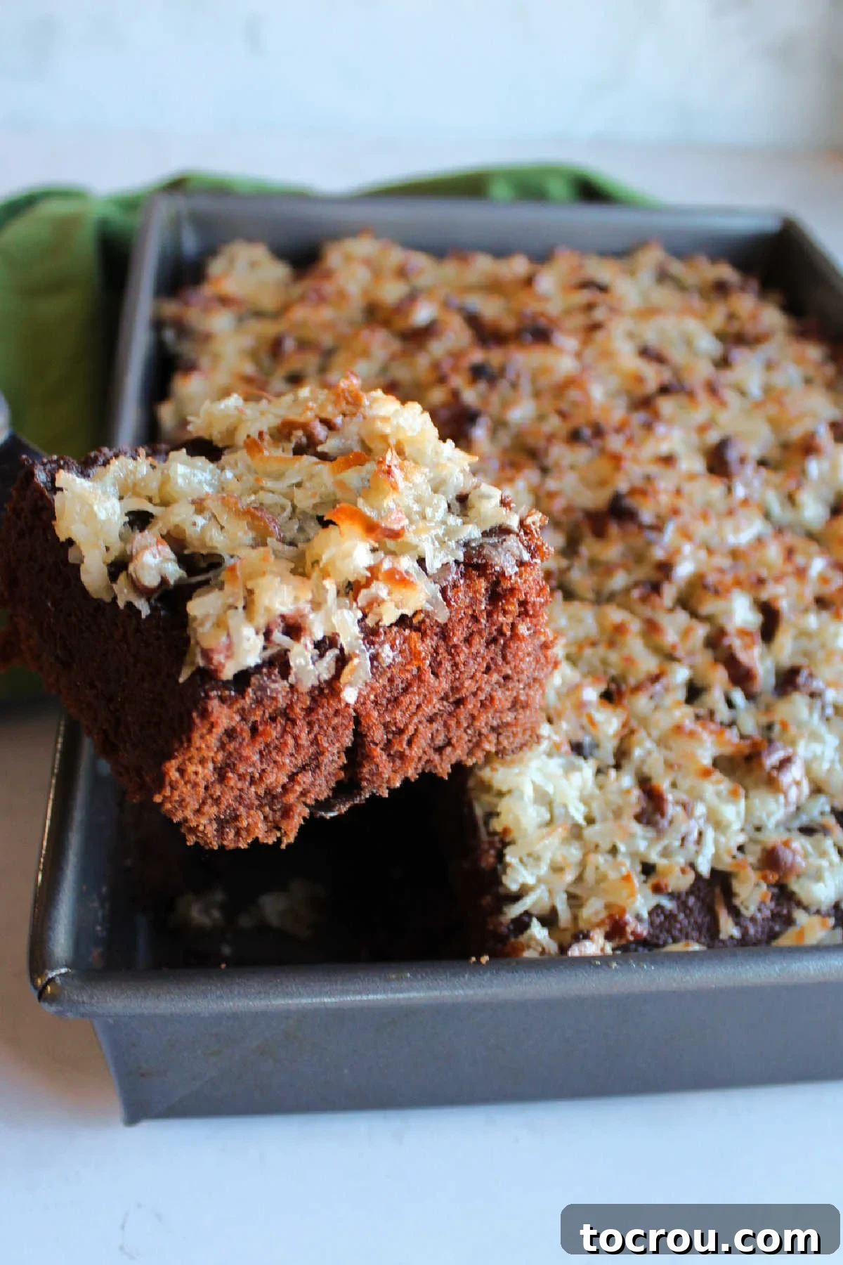 Decadent German Chocolate Cake Crowned with Broiled Coconut 11 A large piece of German chocolate cake with broiled coconut frosting being carefully lifted out of a 9x13-inch baking pan, showcasing its moist texture and golden topping.