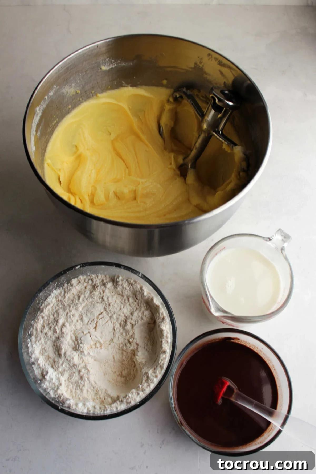 Decadent German Chocolate Cake Crowned with Broiled Coconut 4 A mixing bowl containing a creamy butter, egg, and sugar mixture, flanked by a bowl of dry ingredients (flour, baking soda, salt), a bowl of melted chocolate with water, and a measuring cup of buttermilk.