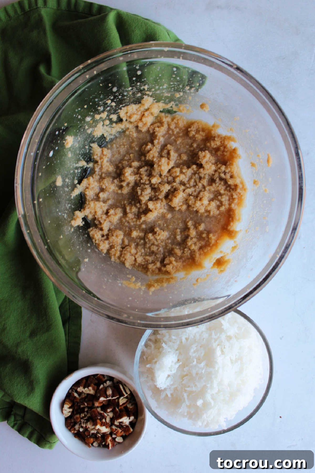 Decadent German Chocolate Cake Crowned with Broiled Coconut 7 A bowl containing a creamy mixture of butter, brown sugar, and milk, positioned next to a bowl of shredded coconut and another bowl of finely chopped pecans, all ready for the cake topping.