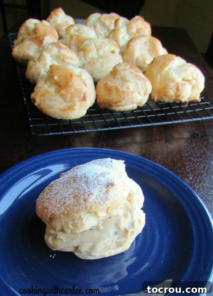A single pumpkin cheesecake cream puff on a white plate, with more empty choux pastry shells visible on a wire rack in the background.