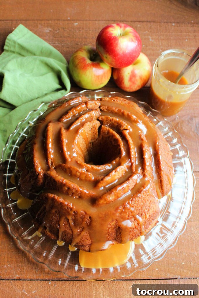 Cider Mill Bundt Cake 5 Whole flower shaped apple cider bundt cake with apples and a jar of caramel sauce in the background.