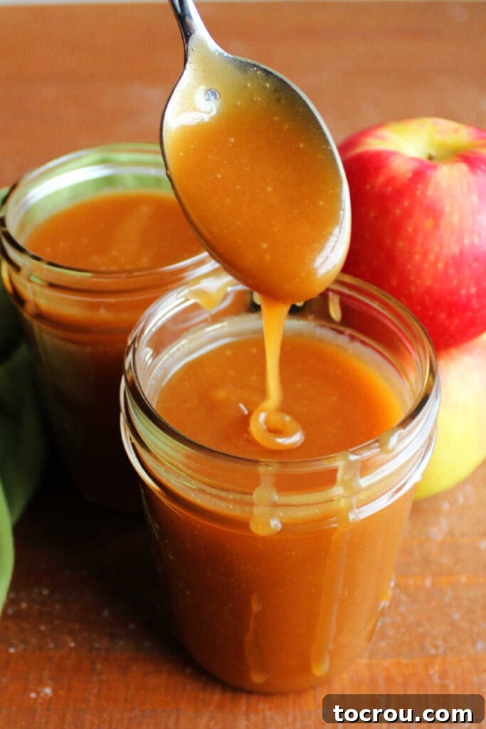 Thick apple cider caramel sauce dripping off a spoon into a jar of more smooth caramel sauce.