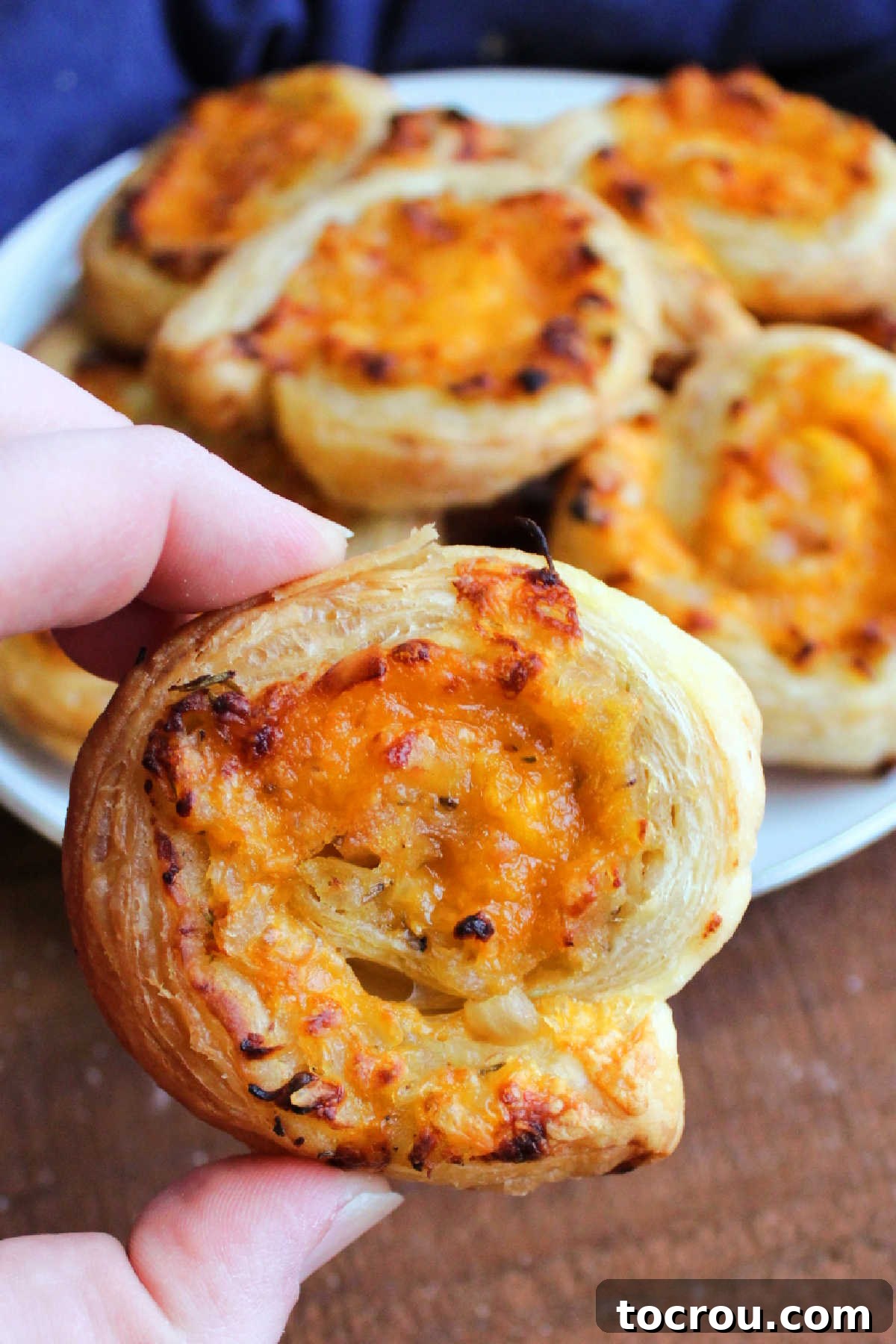 A close-up of a hand holding a perfectly baked apple cheddar puff pastry pinwheel, revealing golden brown pastry and melted cheese with bits of apple.